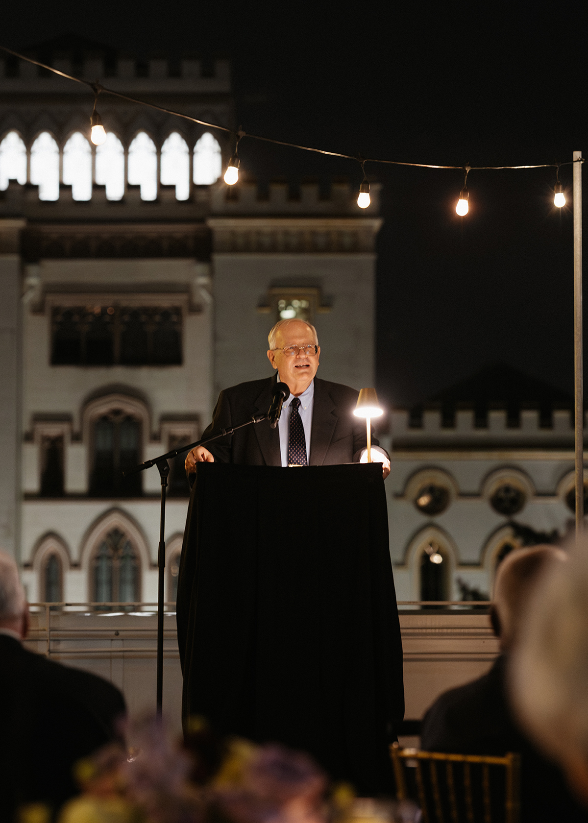 Wearing a dark suit, R. Kelley Pace provides remarks from behind a podium at the Boyd Professor ceremony. This is an outdoor, evening event on a rooftop in Baton Rouge. The lighting is warm and inviting.
