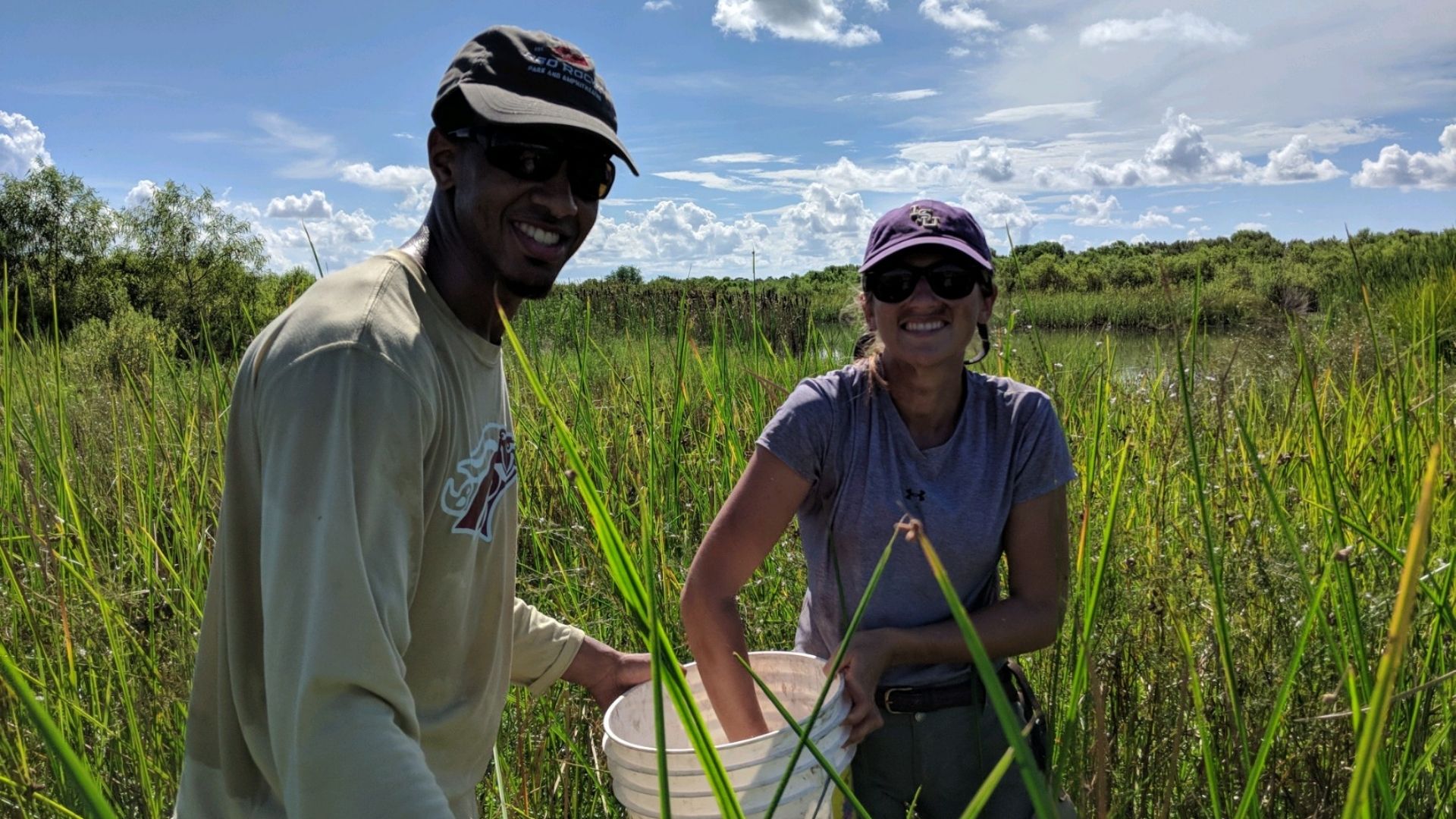 two students in tall grass sticking their hands in a bucket