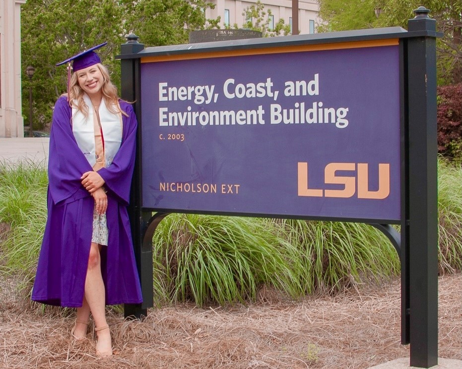 A student in a graduation cap and gown stands by the building sign