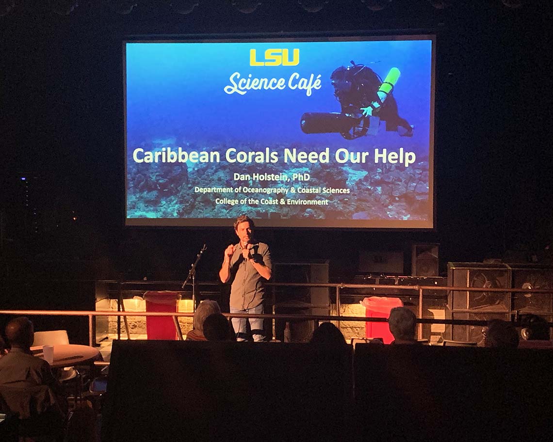 A man with a microphone stands in front of screen saying Carribean Corals need our help