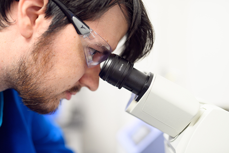 male undergraduate student wearing safety glasses looking into a microscope in Prof. Adam T. Melvin's laboratory