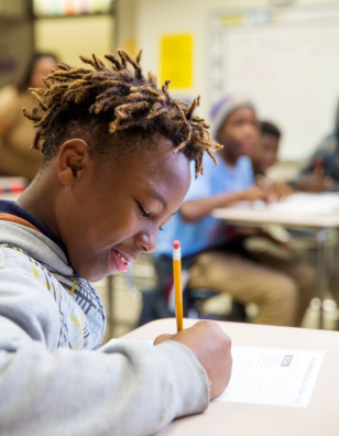 Black male child writing with a pencil and paper in a classroom